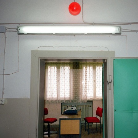 Blick durch eine offene Tür in ein Vernehmungszimmer, in dem zwei rote Stühle einander gegenüber an einem Schreibtisch stehen. Vor dem Fenster hängt eine Spitzengardine.