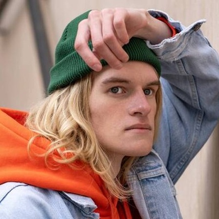 Frustriert schauender Mann mit grüner Mütze, orangen Hoodie und Jeansjacke lehnt an Wand