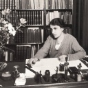 Das Foto zeigt Anna Freud, die jüngste Tochter von Sigmund Freud, in den 1920er-Jahren als junge Frau mit Kurzhaarschnitt. Sie sitzt hinter einem großen Schreibtisch vor einem Bücherregal und blickt nachdenklich schräg am Fotografen vorbei.
