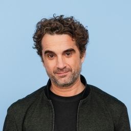 Der Schauspieler Oliver Wnuk trägt ein schwarzes Shirt und eine schwarze Jacke vor blauem Hintergrund und schaut direkt in die Kamera.