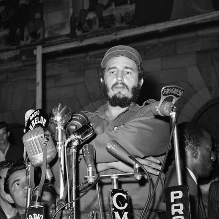 Eine historische scharz-weiß Aufnahme zeigt Fidel Castro angeblitzt vor Mikrofonen am 13. März 1959 vorm Präsidentenpalast in Havanna bei einer Gedenkveranstaltung für die Revolutionäre, die beim Angriff auf den Präsidentenpalast getötet wurden. Der Anschlag wurde von Mitgliedern des Directorio Revolucionario verübt, einer Gruppe, die hauptsächlich aus Universitätsstudenten bestand. Das Directorio arbeitete mit der Bewegung des 26. Juli und anderen revolutionären Gruppen zusammen, um das Batista-Regime zu stürzen.