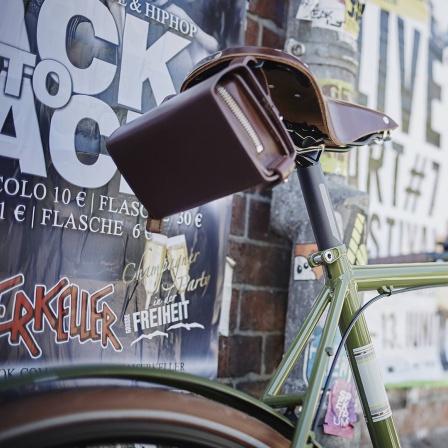 Ein junger Mann steht im Hintergrund in der Unschärfe an eine stark plakatierte Wand gelehnt, im Vordergrund steht ein sehr modernes Fahrrad.