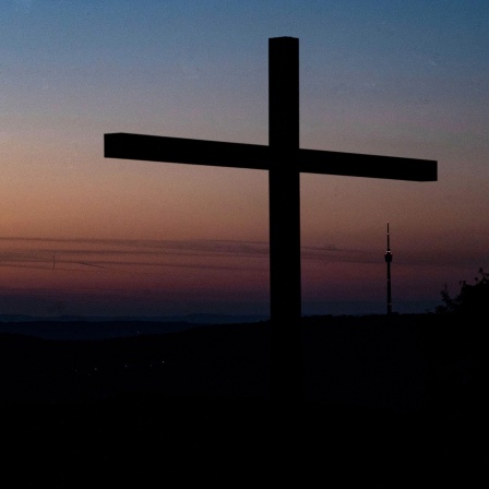 Ein Kreuz steht auf dem Birkenkopf bei Sonnenaufgang vor dem Funkturm und dem Fernsehturm in Stuttgart.