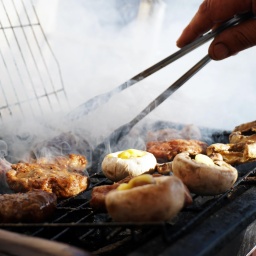 Aus einem dampfenden Grill liegen Gemüse- und Fleischstücke, das gerade mit einer Zange gewendet wird.