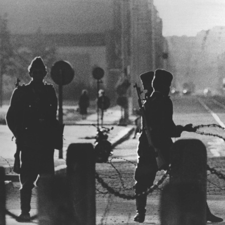 Drei Volkspolizisten ziehen am Potsdamer Platz hinter einer Sperrkette eine Stacheldrahtbarriere. Im Hintergrund ist die menschenleere Leipziger Straße zu sehen. Am frühen Sonntagmorgen des 13. August 1961 wurde unter der Aufsicht von bewaffneten Streitkräften der DDR mit der Errichtung von Straßensperren aus Stacheldraht und dem Bau einer Mauer begonnen