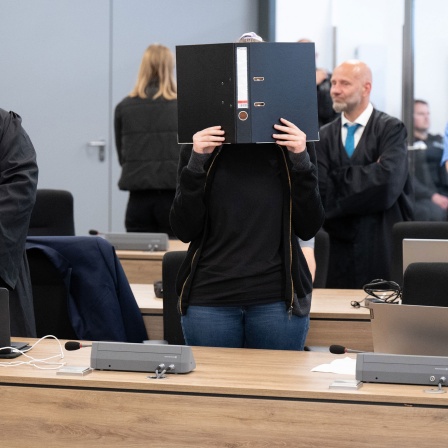 Die Angeklagte (M) steht bei der Fortsetzung des Prozesses im Oberlandesgericht (OLG) Dresden im Verhandlungssaal und hält einen Aktenordner vor ihr Gesicht. 