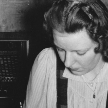 Das Motiv aus der nationalsozialistischen Propaganda zeigt eine Frau beim Schreiben eines Briefes, aufgenommen um 1943/1944. Aufnahmeort unbekannt. Im Hintergrund ein Radiogerät mit dem Foto eines Soldaten darauf stehend.