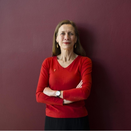 Mariette Rissenbeek steht mit verschränkten Armen in einem roten Oberteil vor einer dunkelroten Wand. 