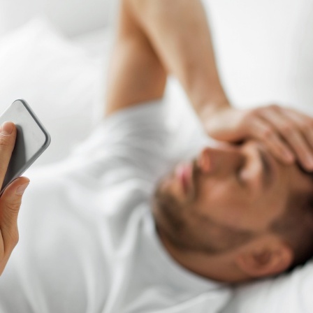 Ein im Bett liegender Mann hält sich mit einer Hand den Kopf, in der anderen ein Smartphone