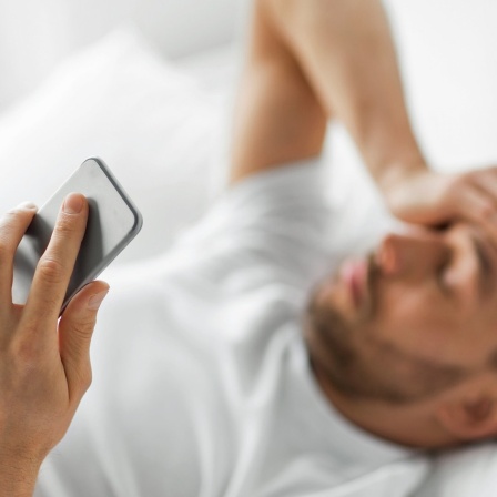 Ein im Bett liegender Mann hält sich mit einer Hand den Kopf, in der anderen ein Smartphone