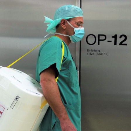 Ein Styropor-Behälter zum Transport von zur Transplantation vorgesehenen Organen wird von einem Mensch in OP-Kleidung am Eingang eines OP-Saales vorbei getragen.