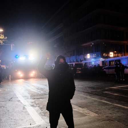 Eine schwarz gekleidete und maskierte Person steht während einer Demonstration im Scheinwerferlicht eines Polizeifahrzeugs und streckt kämpferisch die Faust in die Höhe.