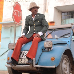 Musiker Roger Damawuzan mit Strohhut und roter Hose sitzt auf einem hellblauen Auto.