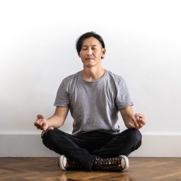Mann in Jeans, T-Shirt und mit Turnschuhen, sitzt im Schneidersitz und meditiert.