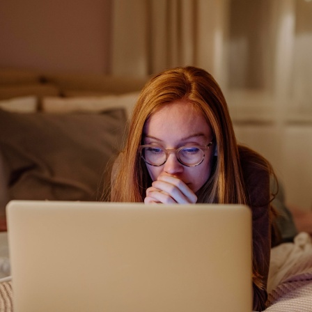 Eine Frau mit Brille liegt in einem Bett und schaut auf den Laptop