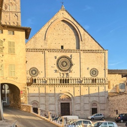 Eine Kirche in Assisi (Italien) im Sonnenlicht, daneben eine Straße und vor der Kirche einige parkende Autos