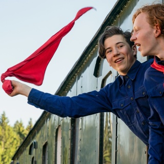 Zwei Jugendliche gucken aus dem Zug und schwenken eine Fahne.