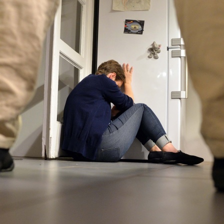 Gestelltes Bild: Eine Person steht vor einer Frau, die sich schützend in eine Zimmerecke drückt.