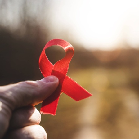 Eine Hand hält eine rote Schleife gegen die Sonne. Symbolbild für Aids und HIV.