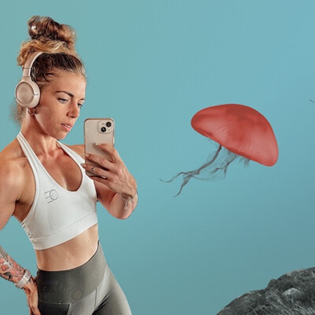 Eine Frau mit Dutt und Kopfhörern steht vor einem Spiegel und macht mit dem Handy ein Selfie. Sie trägt ein Top und hat sehr muskulöse Arme. Im Hintergrund Quallen und Berge.