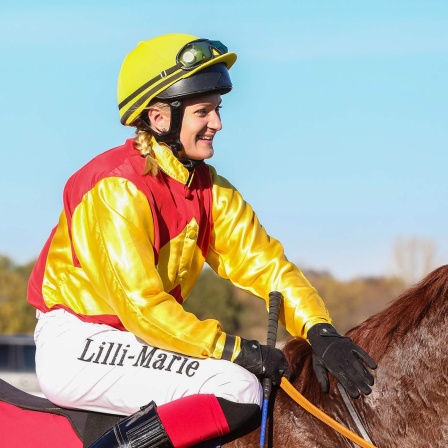 Jockey Lilli Marie Engels sitzt lachend auf dem Rücken eines Pferdes. 