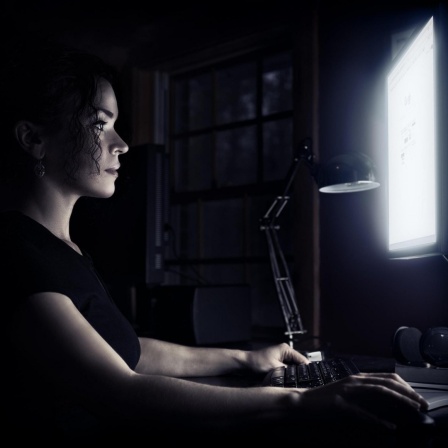 Eine junge Frau sitzt zu Hause an einem Schreibtisch mit einem Computer, ihr Gesicht wird vom Licht des Bildschirms beleuchtet.