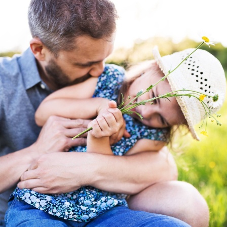 Vater kitzelt kleine Tochter; sie trägt einen weißen Sonnenhut und hält Blumen in der Hand: Kitzeln ist nicht gleich Kitzeln. Psychologen unterschieden schon im 19. Jahrhundert zwei Formen: Knismesis und Gargalesis. Was heißt das?