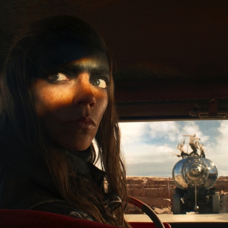 Eine Frau sitzt im dunklen Auto, nur ihre Augen sind hell erleuchtet - im Hintergrund ein Tankwagen - Szene aus "Furiosa"
