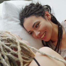 Zwei Frauen liegen gemeinsam in einem Bett und sprechen miteinander, dabei lächelt eine von ihnen, deren Gesicht der Kamera zugewandt ist.