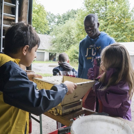 Brighton Paddey Chimanyiwa arbeitet auf dem Schulbauerhof in Bielefeld-Ummeln mit Kindern an der Saftpresse. Im Hintergrund steht ein Pferdeanhänger auf dem Hof, dahinter sind Bäume und Gebüsch zu sehen.