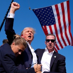 Donald Trump nach dem Anschlagsversuch am 13. Juli 2024 in Butler, Pennsylvania. Er wird von Personenschützern umringt. In seinem Gesicht sind Blutspritzer zu sehen, seine Faust ist gen Himmel gereckt. Im Hintergrund weht die US-Flagge. 