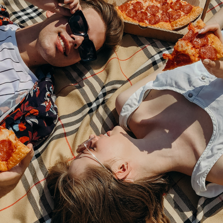 Ein junger Mann und eine junge Frau liegen mit einer Pizza auf der Decke.