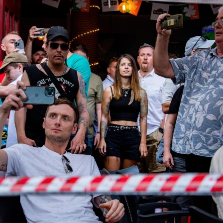 Am Rande eines Protests gegen zu viele Touristen in Barcelona, fotografieren Touristen die Protestierenden, hinter rot-weißem Absperrband.
