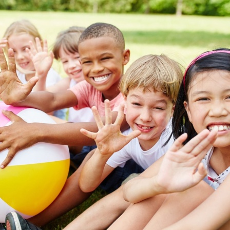 Eine Gruppe multikultureller Kinder sitzen lächelnd und mit einem bunten Ball im Garten im Sommer und winken.