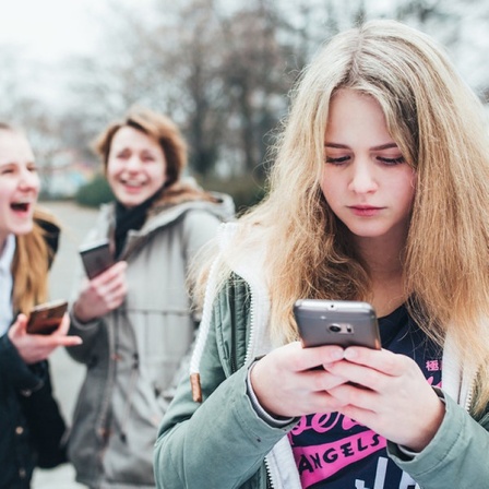 Ein Mädchen schaut auf ihr Handy, im Hintergrund lachen zwei weitere Mädchen.