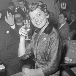Auf dem schwarzweiß Foto sitzt eine lächelnde Frau - Lys Assia - im Kostüm auf einem Sessel und prostet dem Kameramann mit einem Glas Sekt entgegen