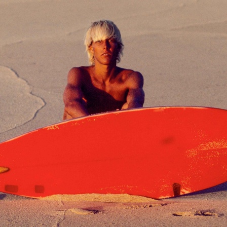 Der amerikanische Surfer Bunker Spreckels sitzt auf einem Strand vor seinem roten Surfbrett.