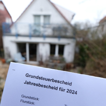 Ein Grundsteuerbescheid für 2024 wird vor einem Privatgrundstück hochgehalten