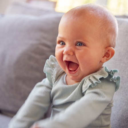 Säugling sitzt auf einer Couch und lacht:  Wenn ein Baby lacht, wenn man es kitzelt - und das tut es schon mit vier Monaten - ist das wirklich schon Humor? Oder nur ein Reflex oder einfach soziales Lachen?