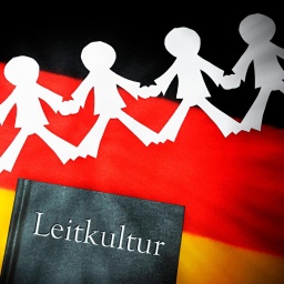 Ein schwarzes Buch mit dem silber gedruckten Wort "Leitkultur" auf einer deutschen Flagge. Über dem Buch liegt ein weißer Scherenschnitt, der Menschen darstellt, die sich an den Händen halten. 
