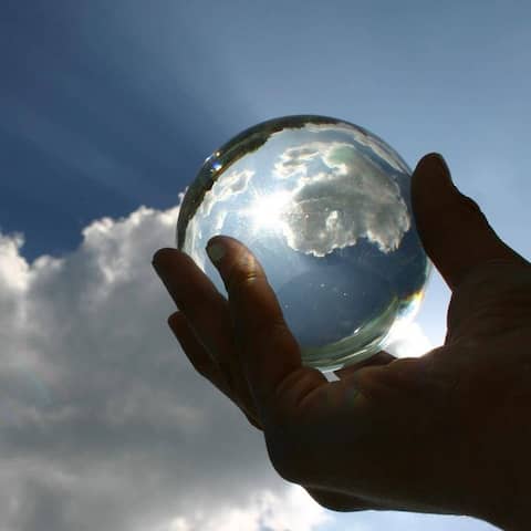 Eine Hand hält eine Glaskugel gegen einen blauen Himmel mit weißen Wolken