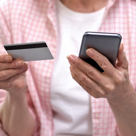 Eine ältere Frau hält eine Kreditkarte in einer und ein Smarphone in der anderen Hand.