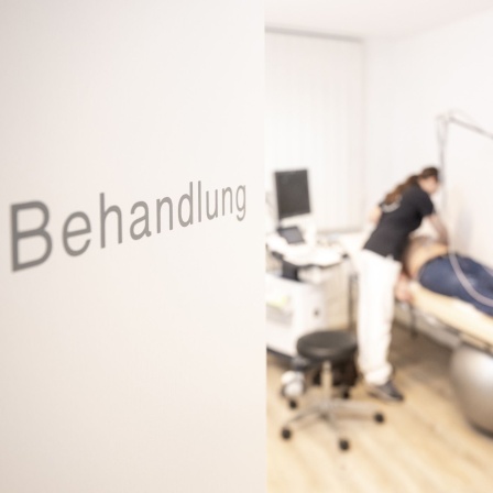 An einer Tür steht "Behandlung", im Hintergrund sieht man einen Patienten auf einer Liege.