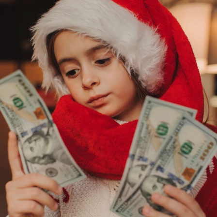 Ein Kind im Schulalter trägt eine Weihnachtsmann-Mütze und Weihnachtsmann-Mantel und blickt auf drei Dollar-Scheine in der Hand.