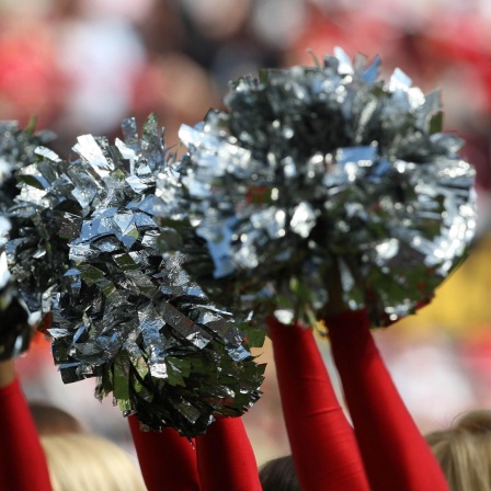 Blick auf mehrere Frauenarme, die silberne Cheerleading-Puschel in die Höhe halten. 