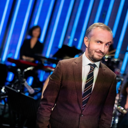 Jan Böhmermann steht im Studio des ZDF Magazin Royal vor seinem Orchester und macht eine ironische Grimasse mit hochgezogener Augenbraue.