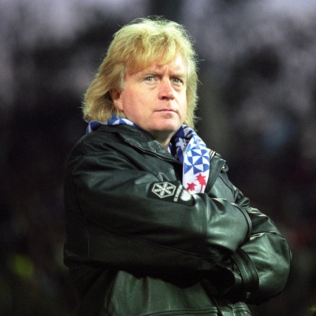 Winnie Schäfer steht mit verschränkten Armen auf dem Platz. Er trägt den blauen KSC-Schal zu einer schwarzen Lederjacke.