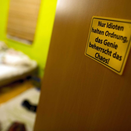 An der Zimmertür des unaufgeräumten Zimmers eines Teenagers klebt ein Schild "Nur Idioten halten Ordnung, das Genie beherrscht das Chaos!" (Bild: picture alliance / ZB)