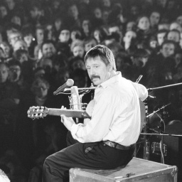 Das Schwarzweißfoto zeigt den Liedermacher Wolf Biermann, aufgenommen seitlich von hinten. Biermann sitzt auf einer Kiste auf der Bühne der Leipziger Messehalle, spielt Gitarre und singt in ein Mikrofon. Im Hintergrund sind zahlreiche Zuschauer zu sehen.