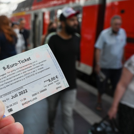 Ein 9-Euro-Ticket wird am Hauptbahnhof hochgehalten, während Zugreisende aus einer Regionalbahn aussteigen.